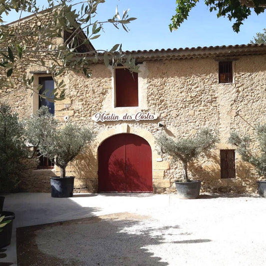 Stone building with a red door and 'Moulin des Costes' sign, surrounded by trees and plants.